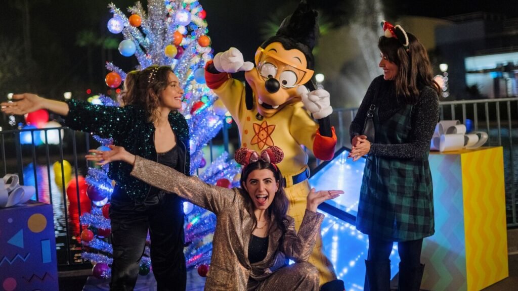 Three women and a Disney character pose and smile near a decorated Christmas tree at night.