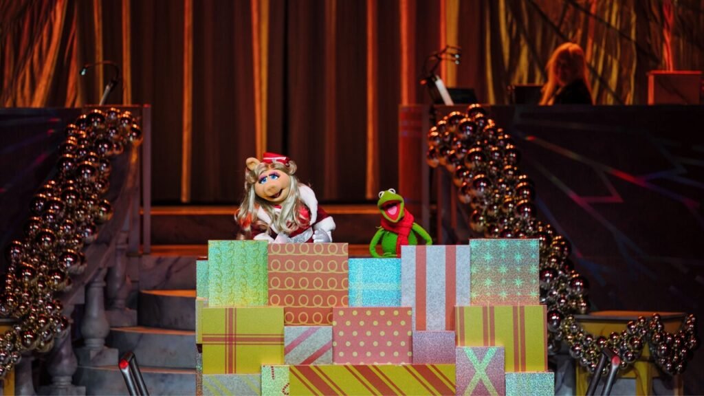Miss Piggy and Kermit the Frog standing behind a stack of colorful gift boxes with a festive background.