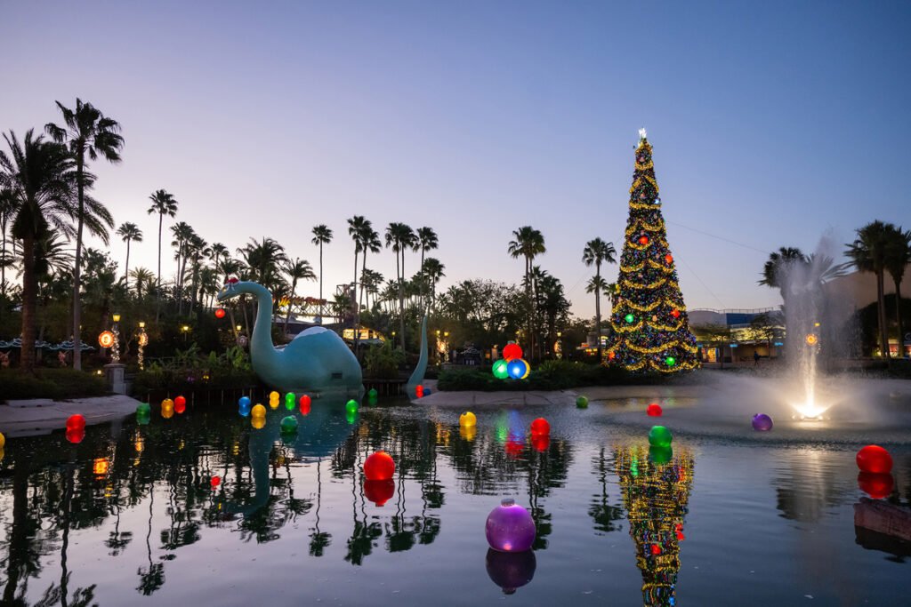 Christmas decorations around a pond with a dinosaur statue, colorful floating ornaments, a decorated Christmas tree, and a fountain at dusk.