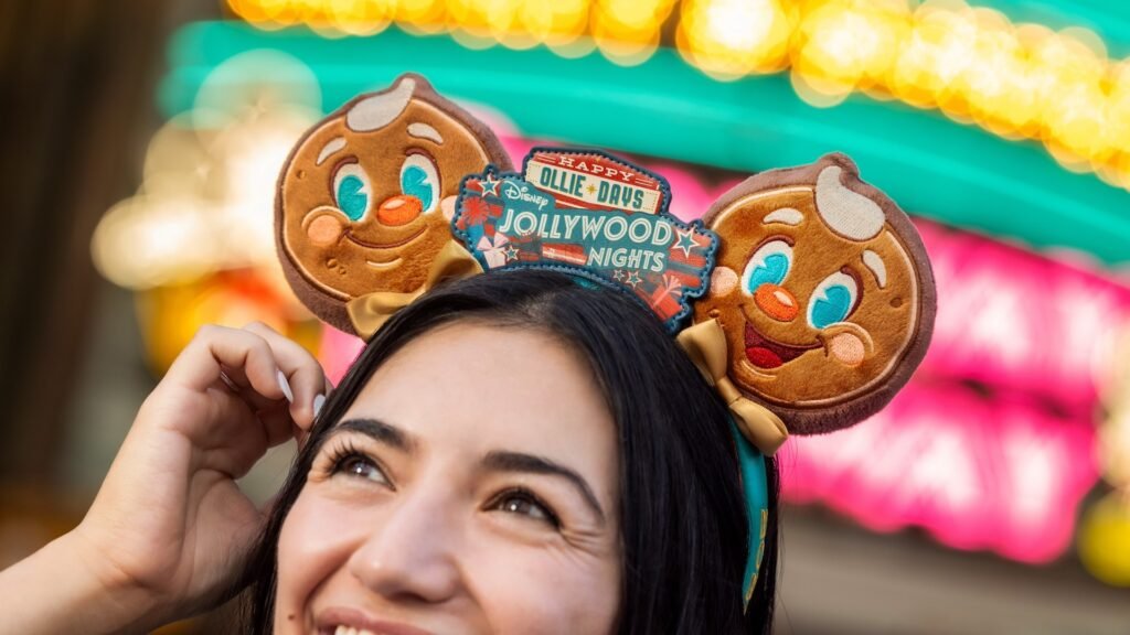 A woman wearing Mickey ear headband featuring gingerbread characters, with a sign reading "Happy Jollywood Nights" on them. A colorful, blurred background of lights emphasizes a festive atmosphere.