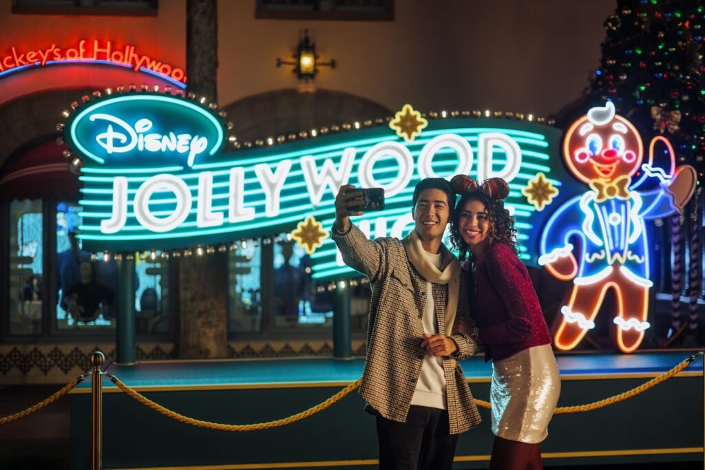 A couple takes a selfie in front of a brightly lit Disney Jollywood Nights sign with neon decorations and a gingerbread character.