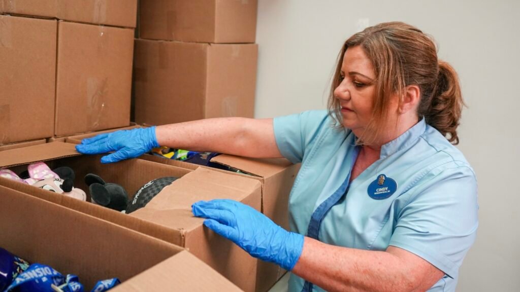 A woman wearing a light blue uniform and blue gloves inspects the contents of a cardboard box filled with toys.
