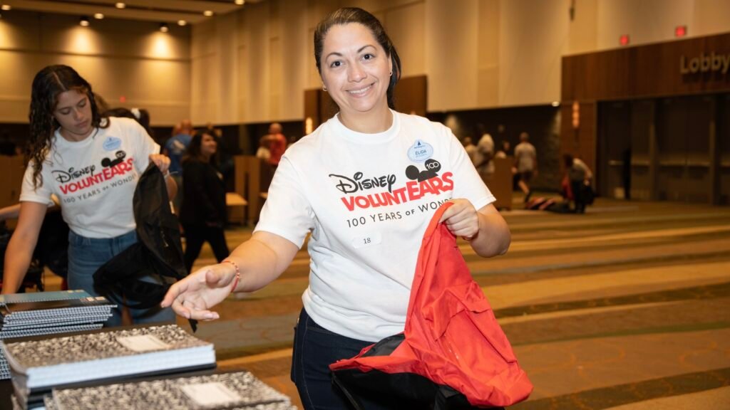 Woman in a "Disney VoluntEARS" T-shirt distributing bags and notebooks at an indoor event.