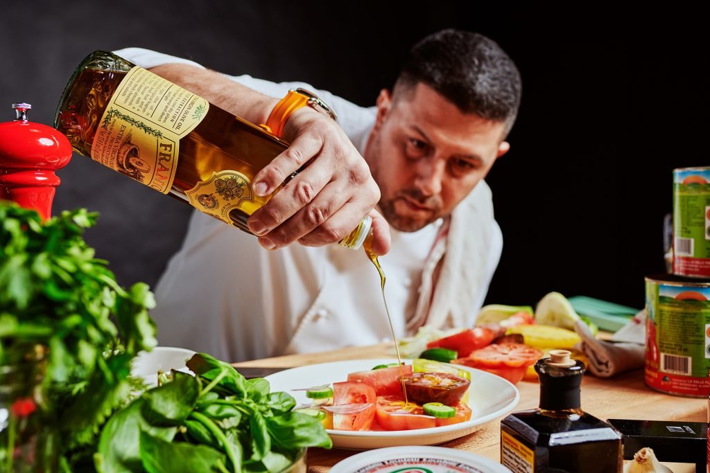 Chef pouring olive oil on sliced tomatoes in a kitchen with various ingredients and canned goods around.