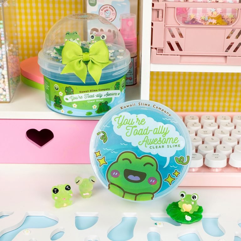 Clear slime container with a smiling frog illustration and the text "You're Toad-ally Awesome." The container is decorated with a green bow and frog charms. Additional cute frog figurines are placed nearby. The scene includes pink and white decor with a heart cutout and keyboard in the background.
