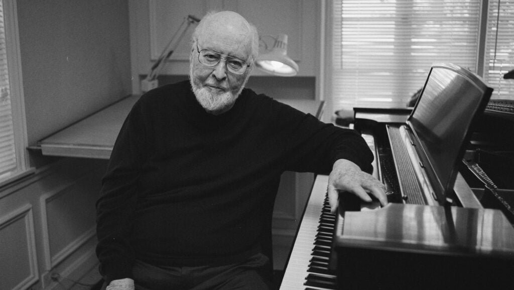 Elderly man with glasses and a beard sitting at a piano, with his hand resting on the piano keys.