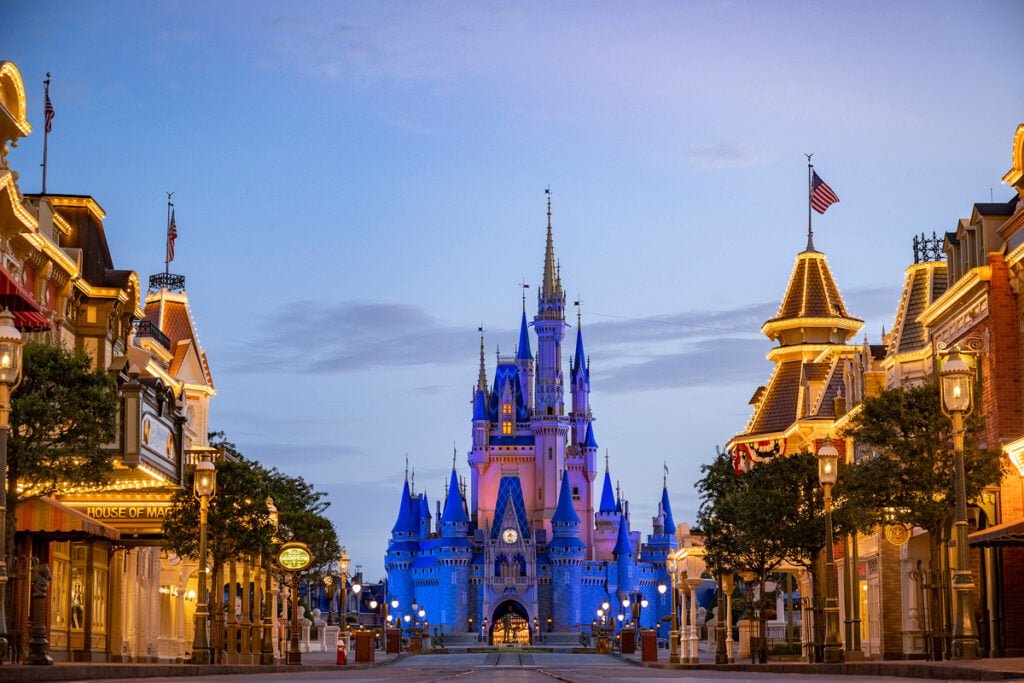 A view of Main Street USA at dawn, leading towards Cinderella Castle at a Disney theme park, with illuminated buildings on either side and an empty street.