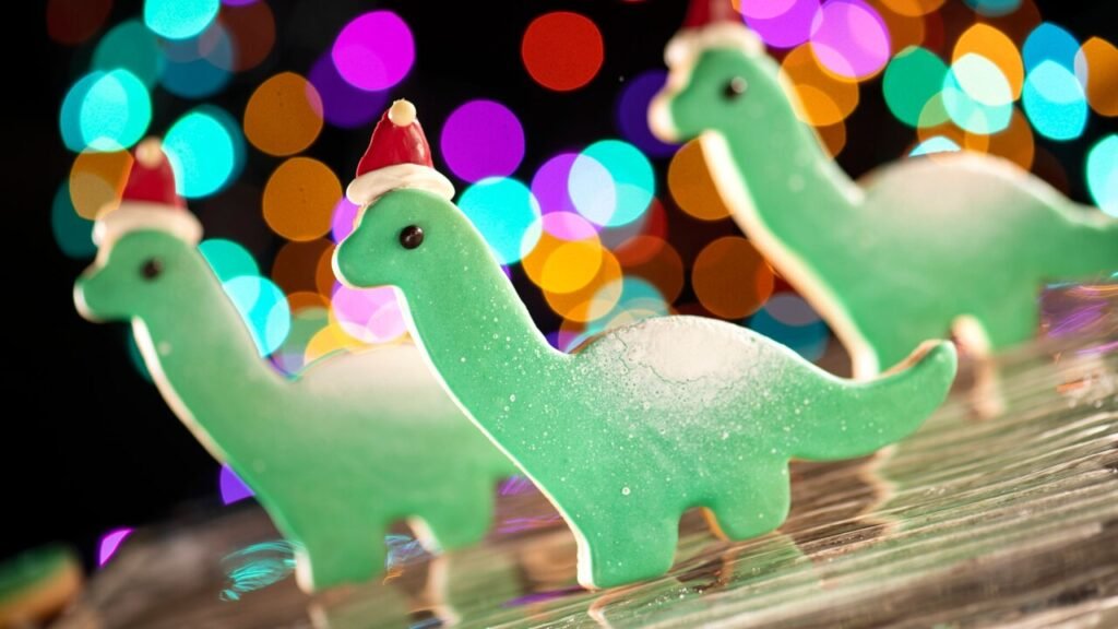 Dinosaur-shaped Christmas cookies with green icing and red Santa hats, against a background of colorful bokeh lights.