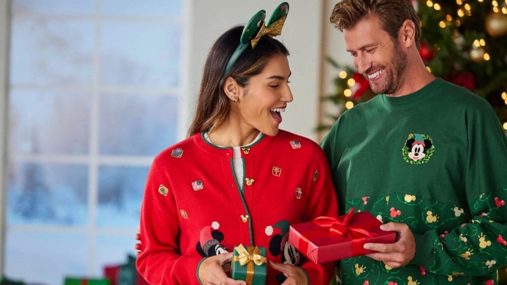A woman in a festive red cardigan with holiday decorations smiles at a man wearing a green sweatshirt with a Mickey Mouse design. They are exchanging gifts in a warmly decorated room with a Christmas tree in the background.