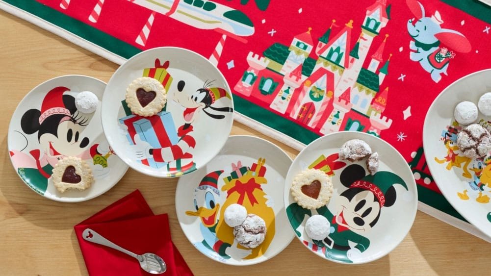 Four decorated plates featuring festive cookies are arranged on a wooden table, accompanied by a red napkin and spoon. The plates depict cartoon characters, including Mickey Mouse and other Disney figures, with a bright holiday-themed table runner in the background.