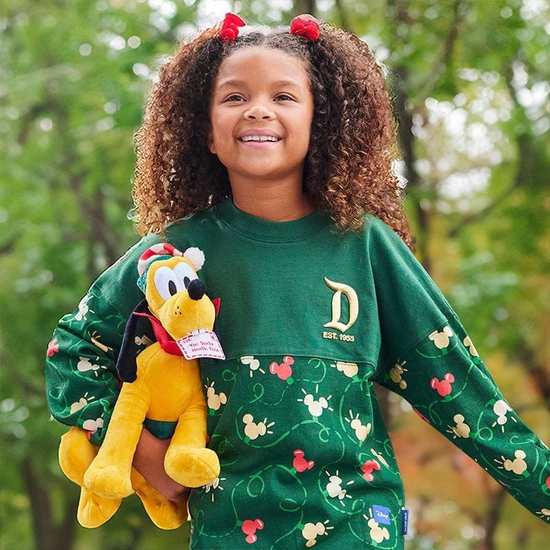 A smiling young girl with curly hair, wearing a green sweatshirt with a Mickey Mouse pattern, holds a yellow plush toy of Pluto against her side.
