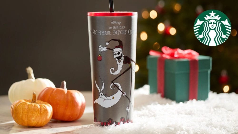 A festive Starbucks cup featuring characters from "The Nightmare Before Christmas," set against a backdrop of pumpkins and a gift box, with a Christmas tree blurred in the background.