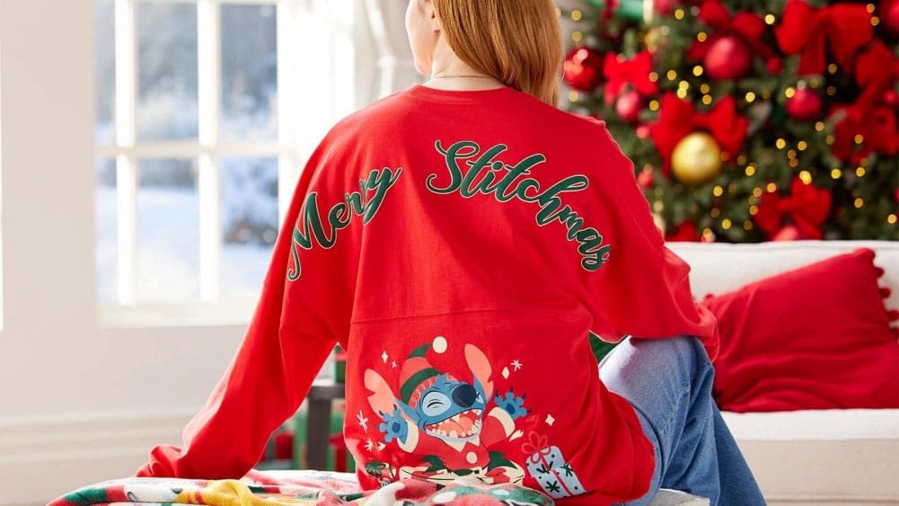 A person sitting on a couch, wearing a red oversized sweatshirt with the text "Merry Stitchmas" on the back, featuring a festive design of Stitch from Lilo & Stitch, in front of a decorated Christmas tree.