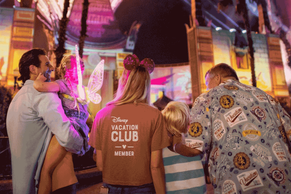 A group of people enjoying an evening at a Disney theme park, with a man holding a child, a woman wearing Mickey Mouse ear headbands and a shirt that reads "Disney Vacation Club Member," and another man in a patterned shirt. The background is brightly lit with colorful structures and palm trees. Disney has announced 2025 DVC Moonlight Magic dates.