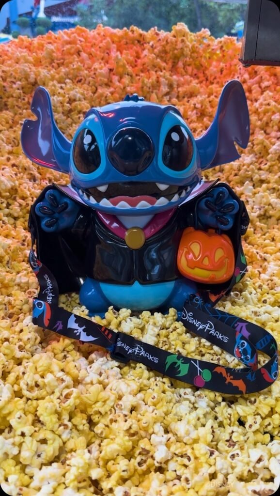 A Vampire Stitch-themed popcorn bucket, designed to resemble Stitch from Disney dressed in a Halloween costume, holding a small pumpkin. The bucket is set against a large backdrop of popcorn.