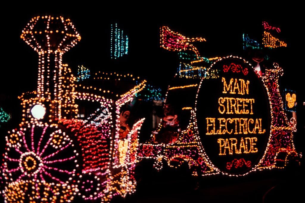 Parade float covered in colorful lights, featuring the words "Main Street Electrical Parade" illuminated in yellow against a dark background.