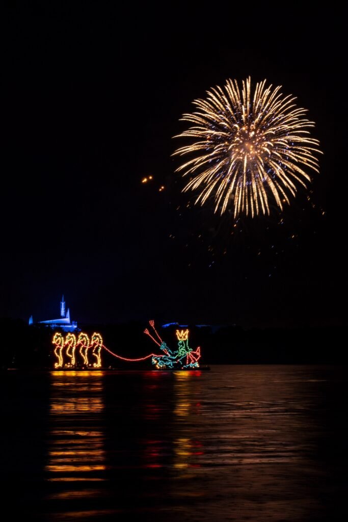 Fireworks bursting in the night sky above a lighted parade float featuring a sea-serpent and a crowned figure on water.