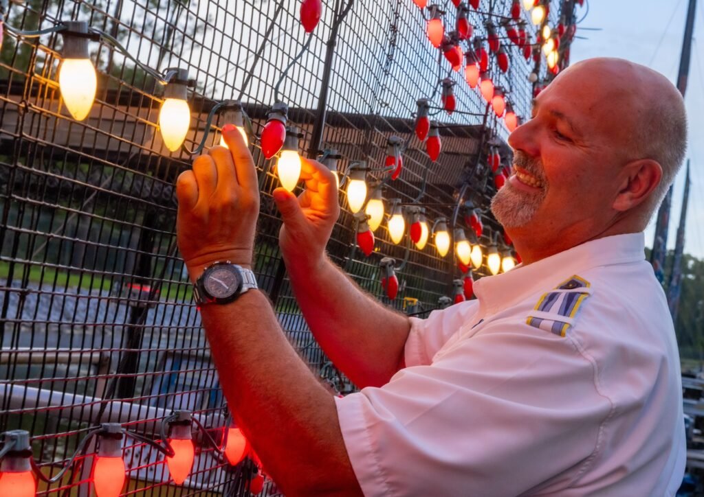 Man in a white uniform adjusts colorful lights on a wire mesh, smiling contently.