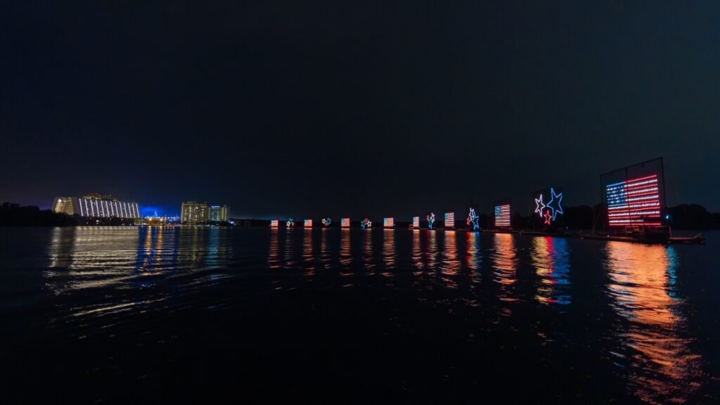 Nighttime view of illuminated buildings and light displays on the water, featuring American flag designs and stars, with reflections on the water.