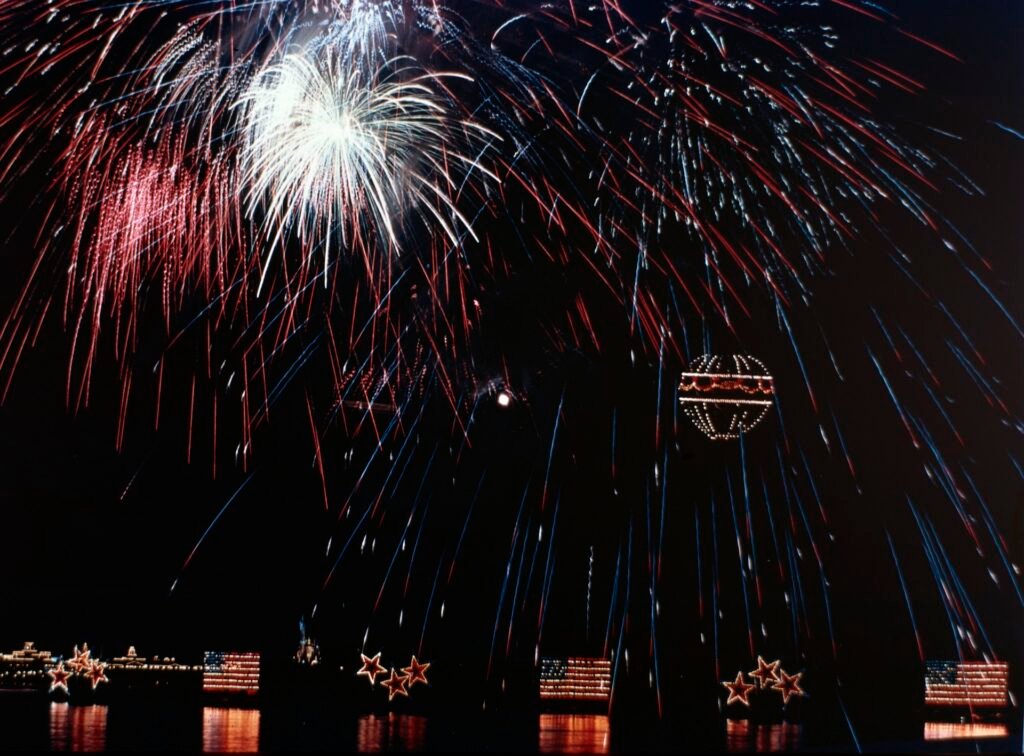 Fireworks bursting in the night sky with illuminated star and flag decorations on water.