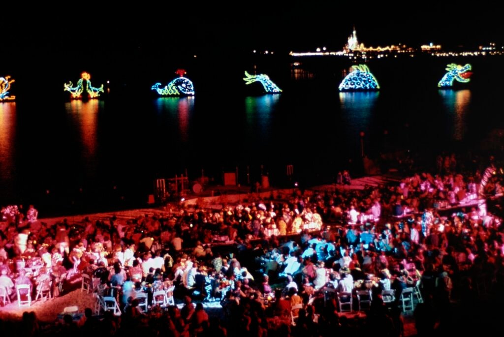 A nighttime celebration by a lake with crowds seated at tables, colorful illuminated sea creature floats in the water, and a distant view of a castle.
