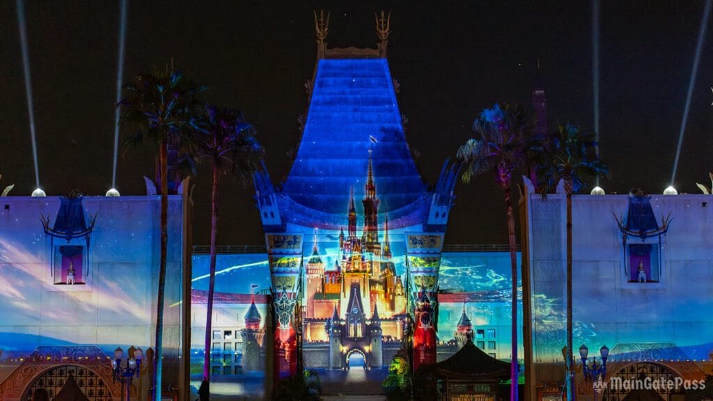 A vibrant light projection of a castle design displayed on a building at night, surrounded by palm trees and illuminated beams of light.