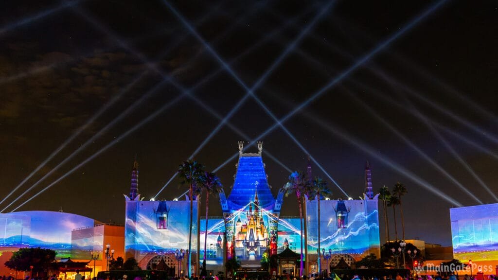 A nighttime view of a themed building illuminated by colorful light projections, with intersecting beams of light spanning the sky and palm trees in the foreground.