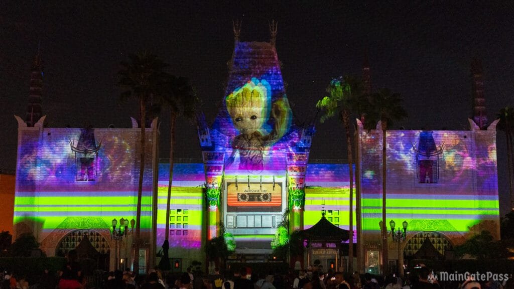 A brightly lit building illuminated with colorful projections, including an image of a character resembling a baby tree, against a night sky. Palm trees and a crowd of people are visible in front of the structure.