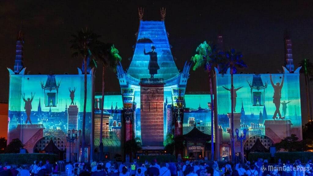 A large building illuminated by colorful light projections at night, featuring silhouettes of people and iconic imagery against a vibrant backdrop. Palm trees are visible in the foreground, and an audience is gathered in front, some holding cameras.