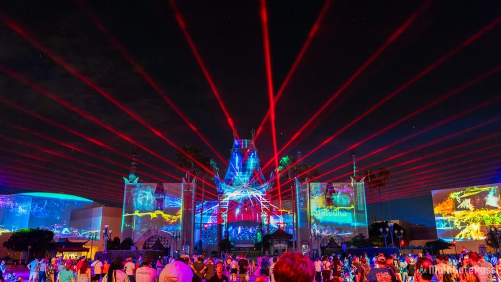 A crowd gathers at night, watching a vibrant light show featuring red laser beams and colorful projections on buildings in a theme park.
