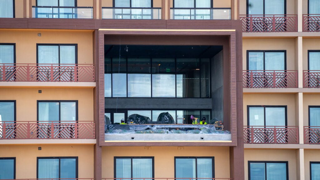 Construction workers in safety gear are working on a large balcony area of a multi-story building, with several large rocks and landscape materials visible. The building features multiple floors with balconies and numerous windows.