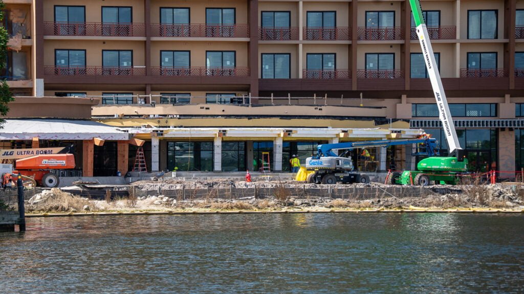 Construction site with two lift vehicles working on a building's exterior, featuring a partially renovated structure with large glass windows, and a view of a water body and rocky shoreline in the foreground.