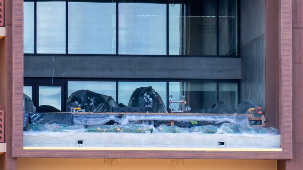 A large window of a building showing a balcony filled with covered plants and gardening tools, with reflections of the sky and surrounding environment in the glass.