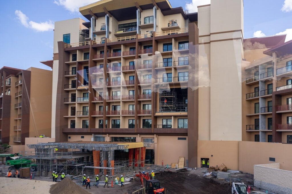 Construction site of a multi-story building with scaffolding and workers in safety vests. The building features balconies and several floors, with some areas under renovation.