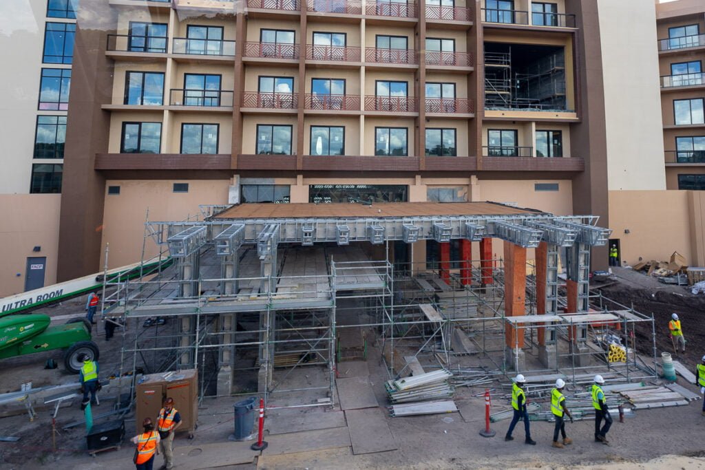 Construction site featuring a building facade with scaffolding and workers in safety gear. A lift is visible, and materials are scattered around the site.