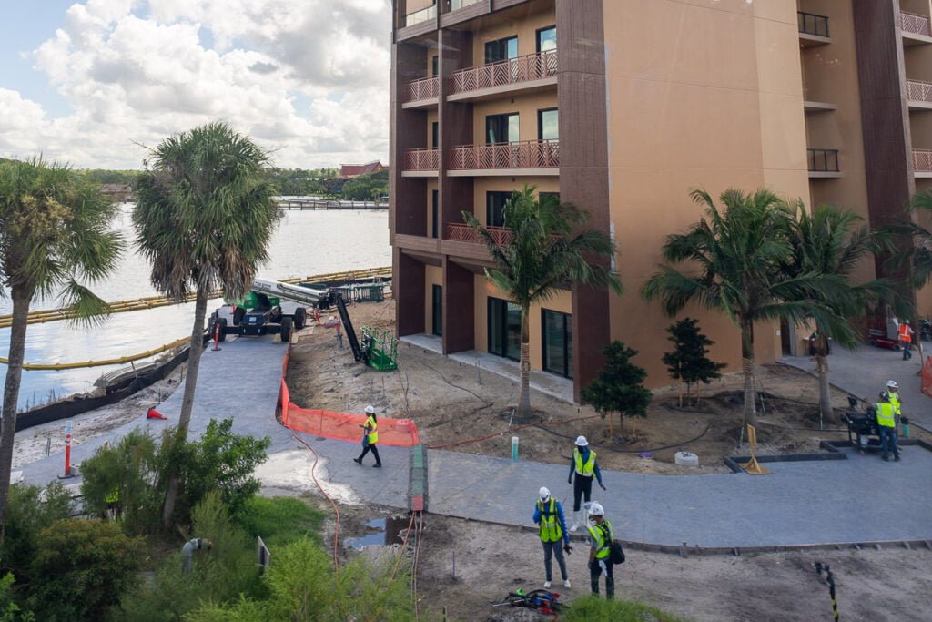 Construction site with workers in safety vests and hard hats, surrounded by palm trees and near a waterway. Heavy machinery is present, and a pathway is being constructed.