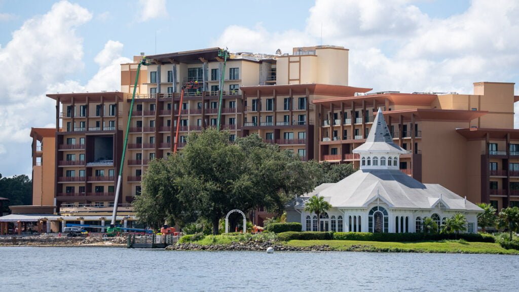 A large hotel building under construction is seen beside a smaller white gazebo structure by the water, with trees and greenery in the foreground.