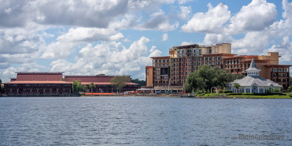 A scenic view of a lakeside resort featuring a large multi-story building, a lower building with a red roof, and a white gazebo in a lush green area, all set against a backdrop of fluffy clouds and blue sky.