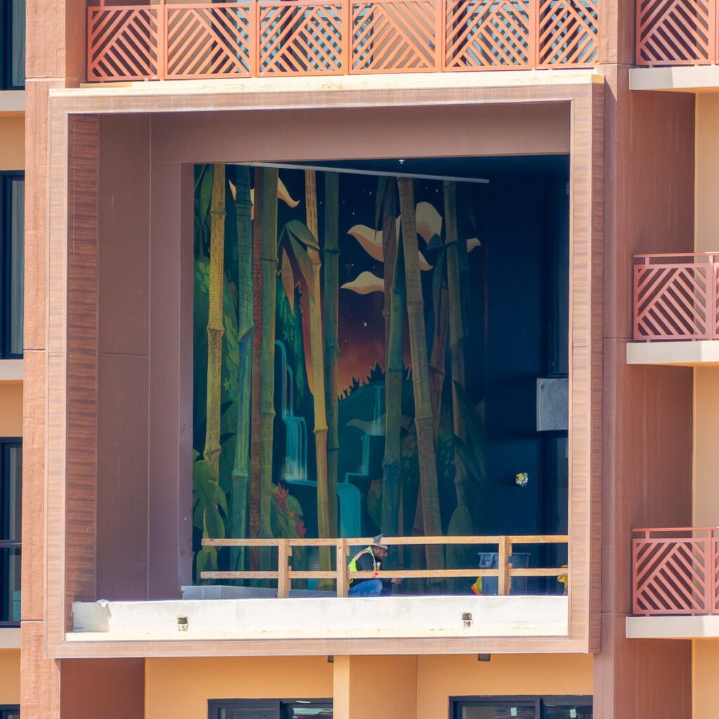 A worker in a safety vest is seen sitting on a ledge in front of a large mural depicting a forest with bamboo and waterfalls, illuminated by a sunset sky. The mural is located on the exterior wall of a building, partially framed by a balcony railing.