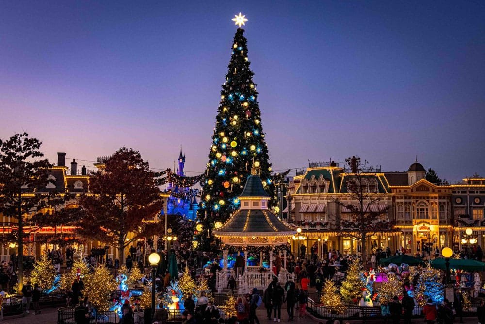 A beautifully decorated Disneyland Paris Christmas tree with colorful ornaments and a star on top stands in the center of a festive plaza, surrounded by twinkling lights, snowman figures, and holiday decorations, as people gather to enjoy the holiday atmosphere at dusk.