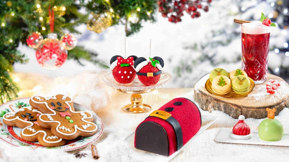 A festive display of Christmas-themed desserts including gingerbread cookies, decorated strawberries resembling Santa, a red and black cake shaped like Santa's mailbox, colorful macarons with holiday designs, and a red beverage beside various decorative treats. Snow and a decorated Christmas tree provide a seasonal backdrop.