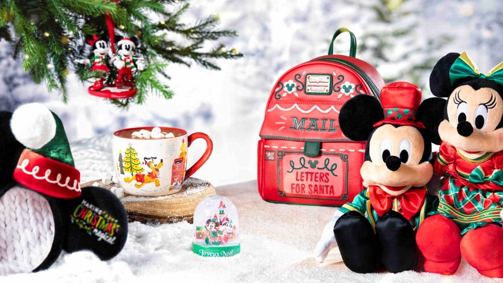 A festive scene featuring Mickey and Minnie Mouse plush toys, a red backpack labeled "MAIL" with "LETTERS FOR SANTA," a mug filled with hot chocolate and marshmallows, a decorative snow globe, and holiday-themed accents under a Christmas tree with ornaments.