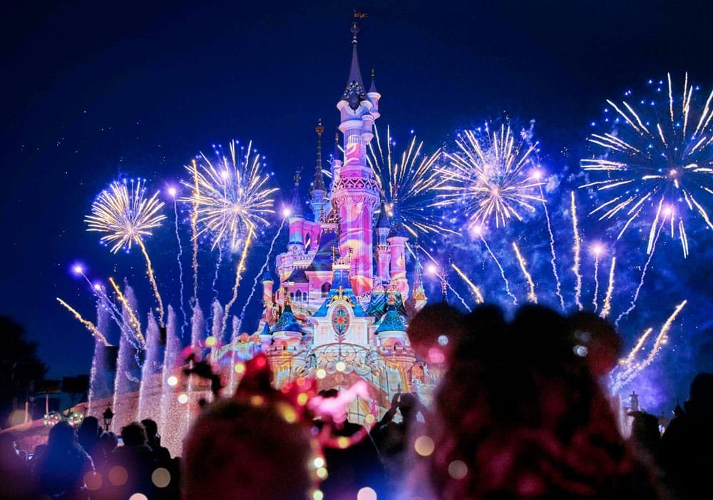 Colorful Disneyland Paris Christmas fireworks illuminate a castle at night while a crowd watches in the foreground, with some people wearing Mickey Mouse ears.