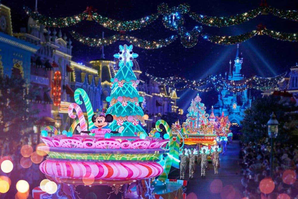 A vibrant Disneyland Paris nighttime parade featuring colorful floats decorated for the holidays, with a prominent Christmas tree and a character in festive attire. The street is lined with twinkling lights and festive decorations, and a castle is faintly visible in the background.
