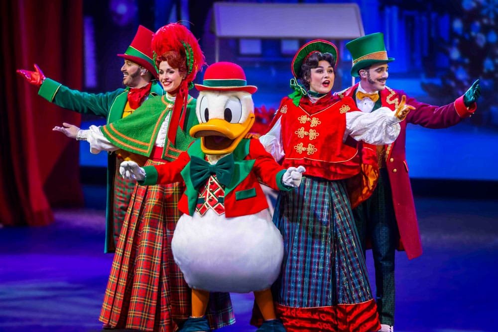 A group of performers in festive costumes, including a large character resembling a duck, are smiling and posing on stage. The backdrop suggests a winter or holiday theme.