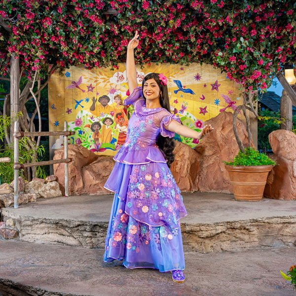 An Encanto character, a woman in a colorful floral dress poses with one arm raised on an outdoor stage decorated with flowers and a mural in the background of a meet and greet.