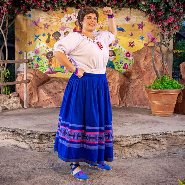 An Encanto character wearing a white blouse, blue skirt with colorful designs, and blue shoes is posing with a confident, flexed arm stance in front of a colorful background for a meet and greet.
