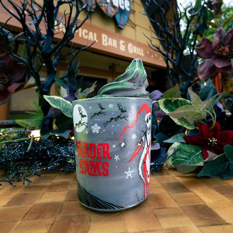 A decorative cup featuring Halloween and winter themes, displaying a character resembling Jack Skellington with a festive hat, surrounded by dark foliage and a bar sign in the background.