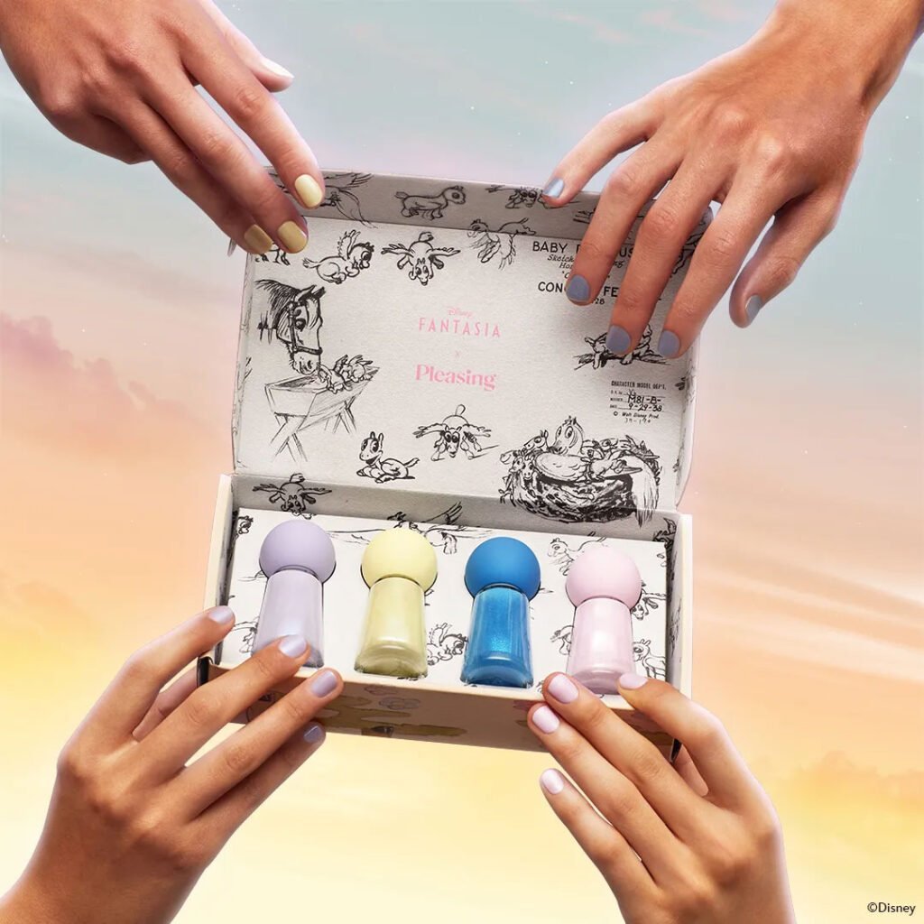 A close-up of two hands holding a box featuring colorful nail polish bottles, with a pastel sky background and cartoon illustrations inside the box. The bottles are in purple, yellow, blue, and pink, while the hands have various nail polish colors applied.