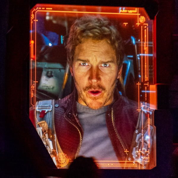 A man with short, tousled hair and a slight beard is looking intently at the camera, with an expression of surprise or concern. He is dressed in a dark jacket over a light shirt, and the image is framed with a futuristic digital overlay, suggesting a sci-fi context.